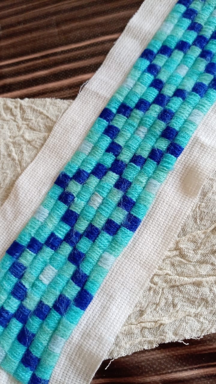 Pulsera tejida con patrón geométrico en turquesa y azul marino sobre banda de tela blanca, detalle del proceso de confección